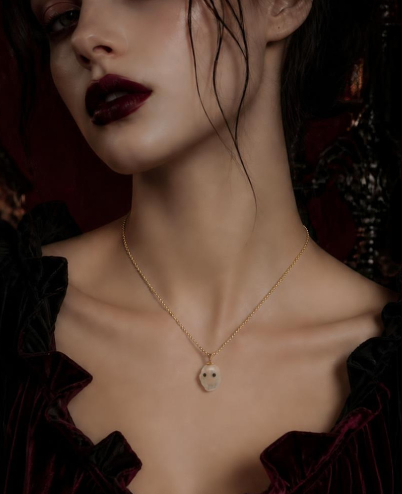 A close-up portrait of a woman wearing a delicate gold necklace with a small skull pendant, styled in a dark, gothic setting with candles and deep red velvet clothing.